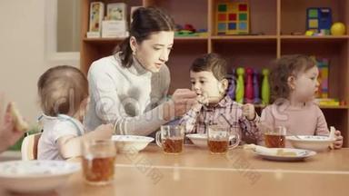 幼儿园老师和孩子们一起坐在桌子旁，用<strong>勺子</strong>喂那个黑发小男孩，另<strong>一个</strong>
